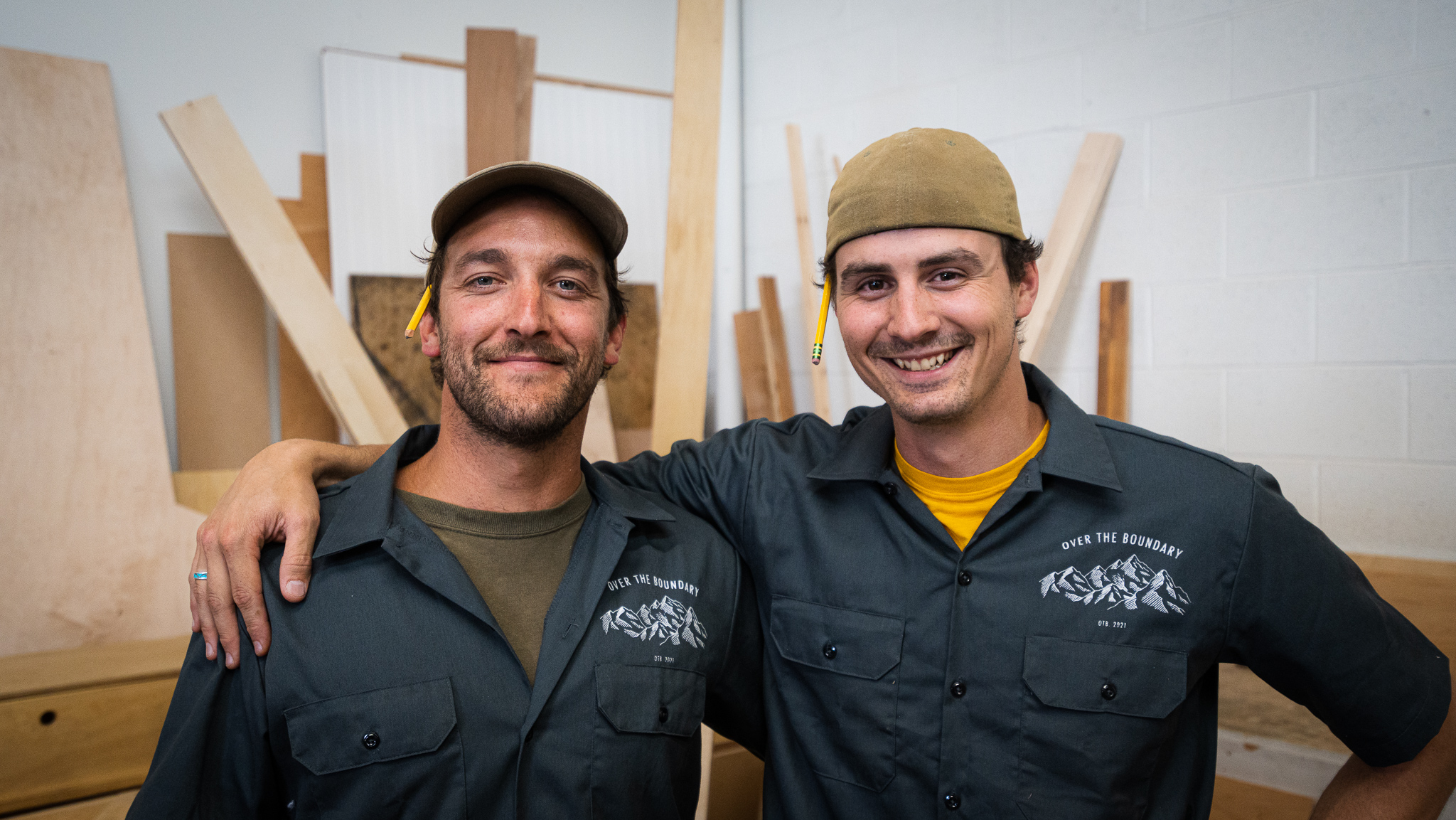 A picture of of Will and Buzz, the co-owners of OTB Fabrication, with their arms around each other smiling for the camera. They're both wearing OTB Fabrication shop shirts, and wood can be seen stacked in the background.